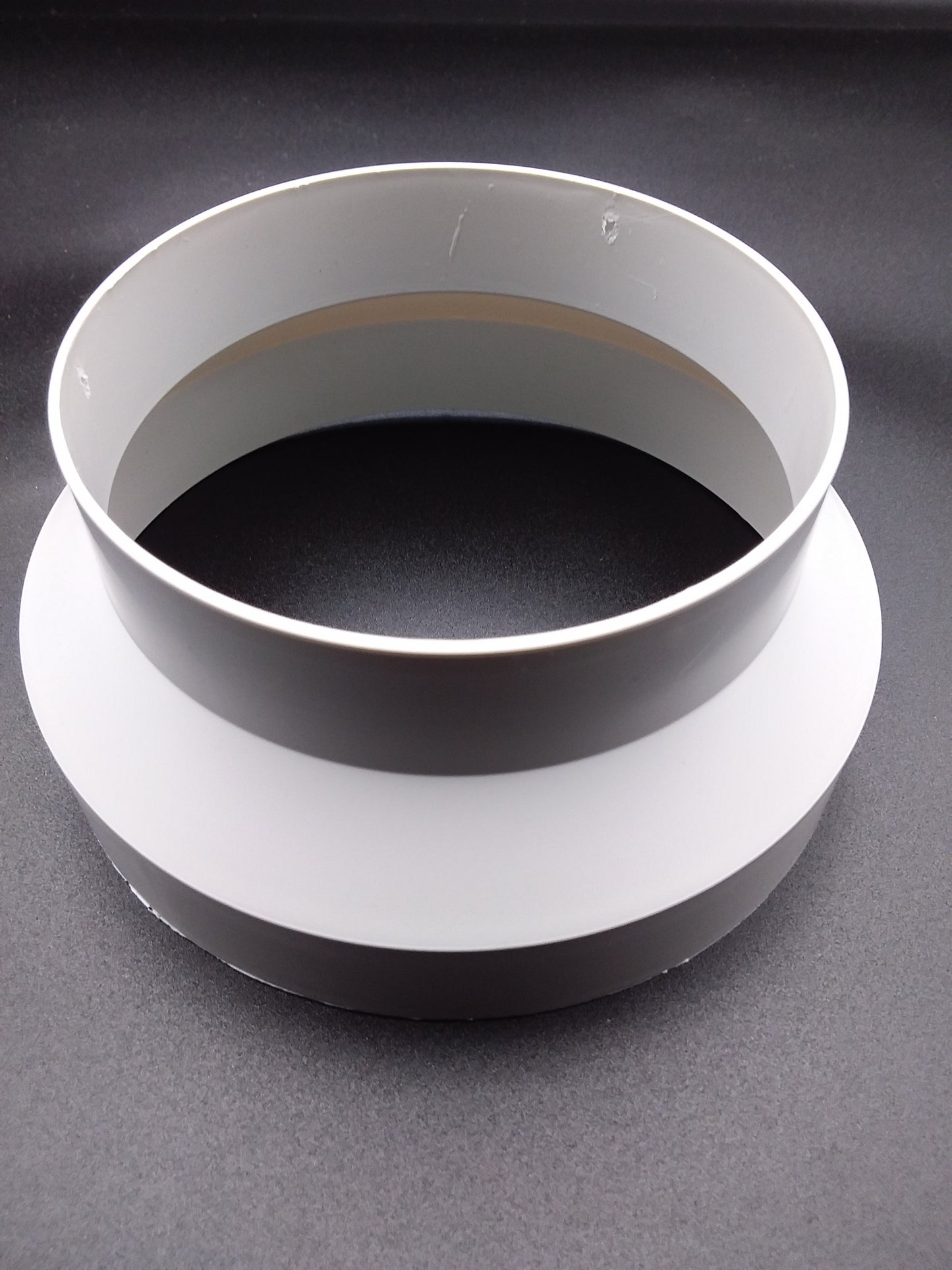 A white ducting reducer, with a round shape and two concentric rings, placed on a flat surface.
